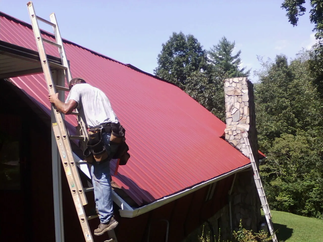 Roofer on a ladder beside a red metal roof and stone chimney — Soffit & Fascia Repair by Roofing Brothers in Mason